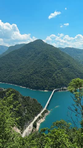 Scenic view of turquoise Piva Lake winding between steep forest-covered mountains in Montenegro. A narrow mountain road follows the shoreline through the dramatic green landscape.