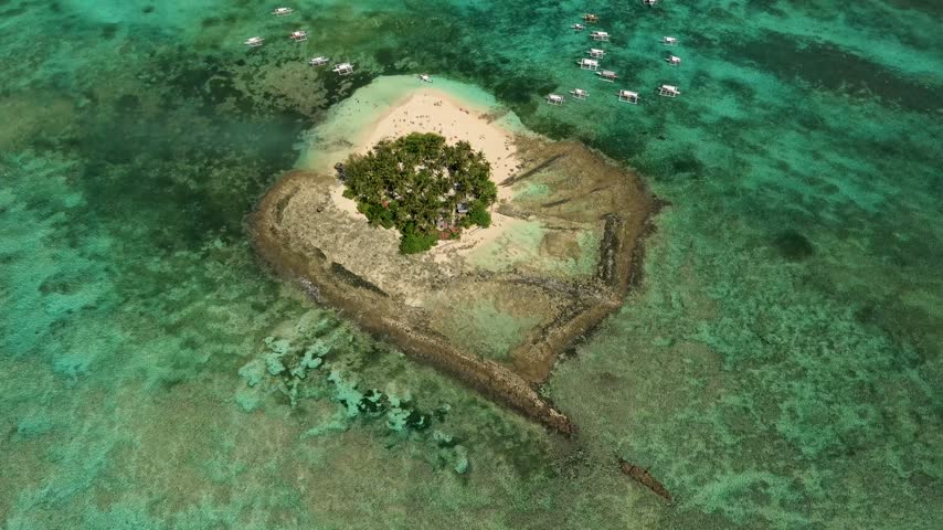 Aerial view of the distinctive Guyam Island in Siargao, Philippines, surrounded by extensive shallow coral reefs and turquoise ocean waters, with a small white sand beach and palm-covered interior.