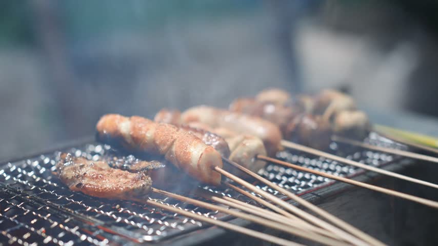 Grilled skewers of chicken, sausage, and meatballs sizzling on a metal grill over glowing coals at an outdoor street food stall