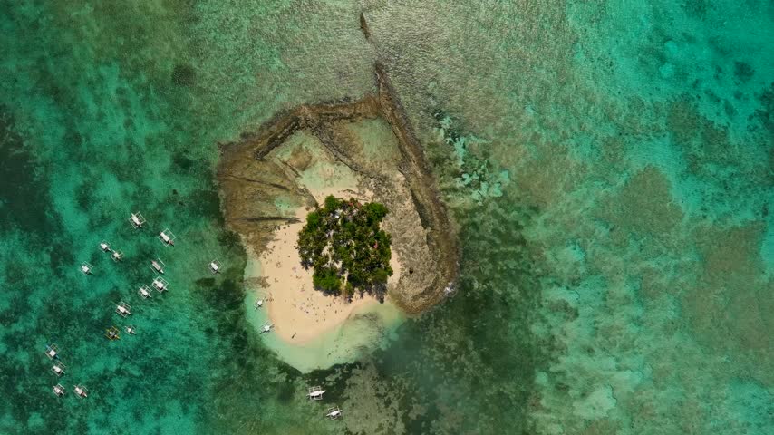 Aerial view of the distinctive Guyam Island in Siargao, Philippines, surrounded by extensive shallow coral reefs and turquoise ocean waters, with a small white sand beach and palm-covered interior.