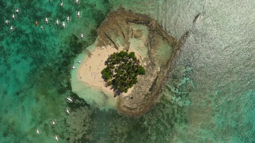 Aerial view of the distinctive Guyam Island in Siargao, Philippines, surrounded by extensive shallow coral reefs and turquoise ocean waters, with a small white sand beach and palm-covered interior.