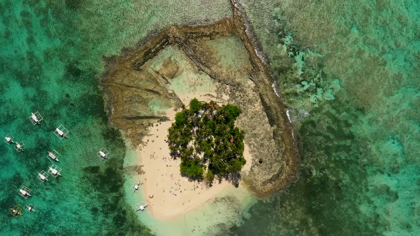 Aerial view of the distinctive Guyam Island in Siargao, Philippines, surrounded by extensive shallow coral reefs and turquoise ocean waters, with a small white sand beach and palm-covered interior.