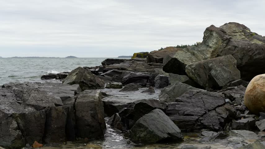 Rocky coastline with gentle ocean waves splashing against dark, jagged rocks along Englishman Bay at Roque Bluffs State Park in Washington County, Maine.