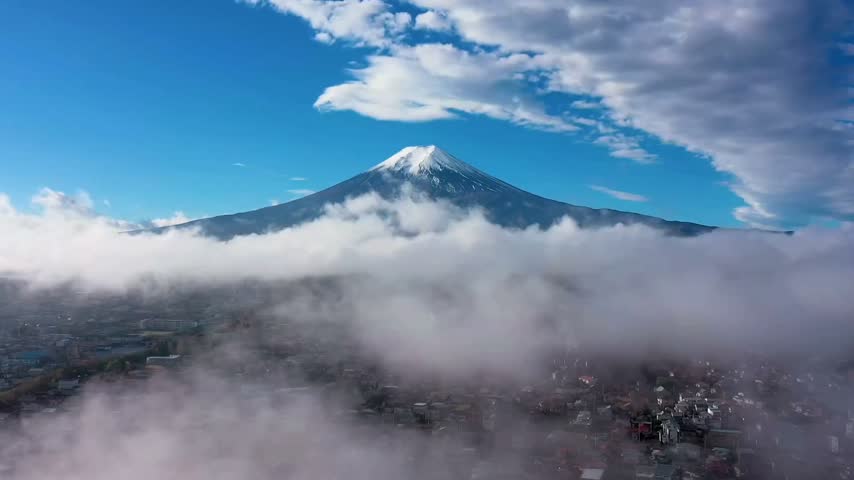 Cinematic aerial drone footage of snow-capped Mount Fuji rising above a sea of clouds, with the surrounding landscape partially visible beneath the mist. Beautiful natural mountain scenery, dramatic sky, and peaceful atmospheric landscape suitable for travel, nature, tourism, geography, and scenic background content.
