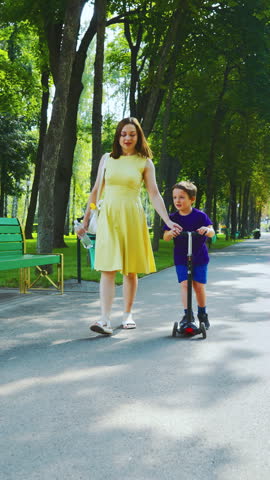 Young woman in yellow dress holding water bottle and petting smiling boy. Loving family members enjoying summer day on sunny park path