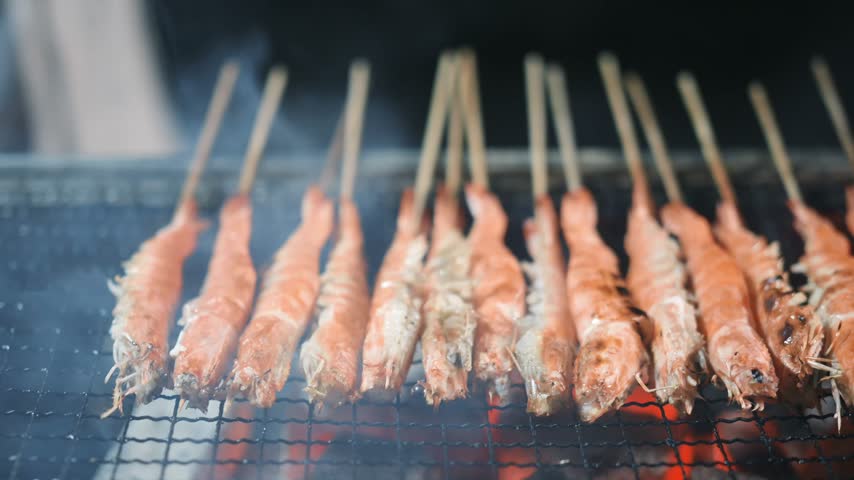 Grilled shrimp skewers sizzling over glowing charcoal on a metal grill at a night food market