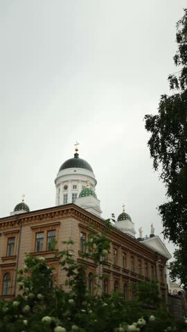 Vertical screen low angle view of a historic building and the white domes of Helsinki Cathedral in Finland with old city architecture green trees and a cloudy sky
