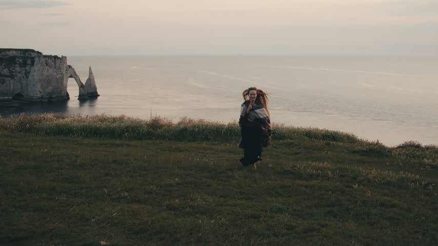 Beautiful young blonde woman in elegant black dress walking running and standing by the sea in Etretat France with long hair flowing in the wind grass cliffs ocean and romantic peaceful coastal