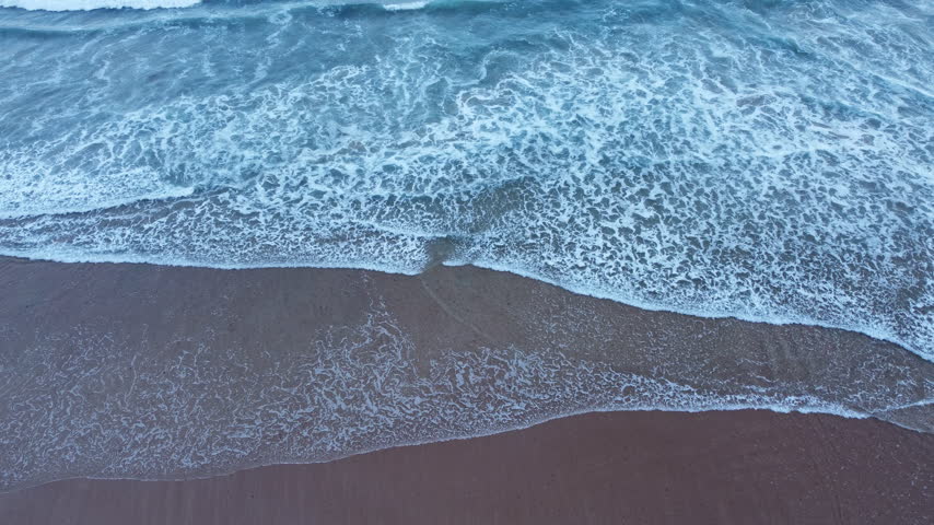 Aerial view of rhythmic waves reaching a dark sandy beach, creating soft white foam, layered shoreline lines, and a peaceful coastal scene.