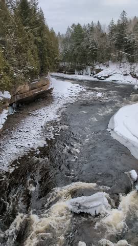 Partially frozen river flowing through a snowy, forested landscape under an overcast sky, Canada
