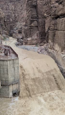 Concrete dam structure on the left, with muddy, fast-flowing water cascading over its edge and continuing downstream through a narrow canyon