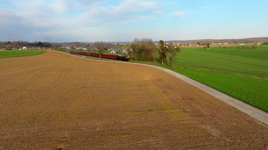 A train moves steadily along its tracks through open fields and rolling hills. The landscape features patches of green grass and harvested brown fields under a blue sky.