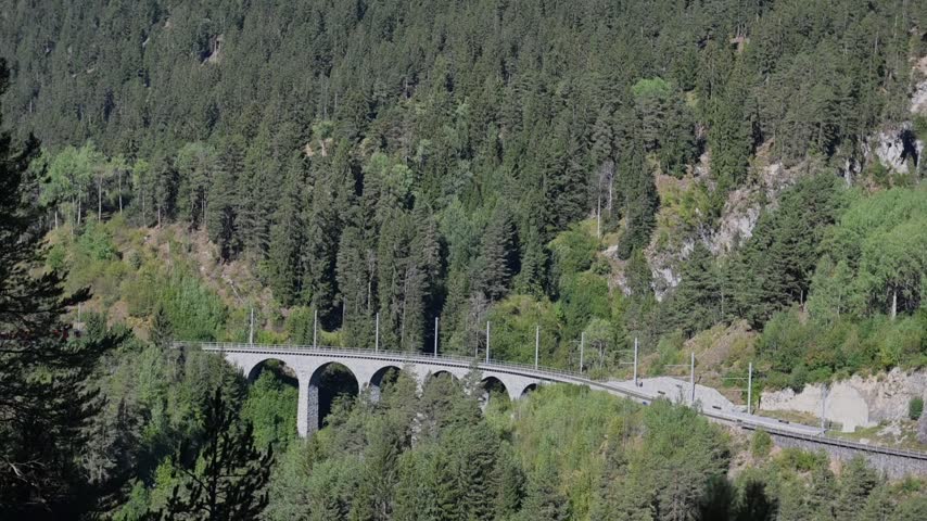 Scenic red passenger train crossing the historic Landwasser Viaduct through a lush alpine forest near Davos, Switzerland.