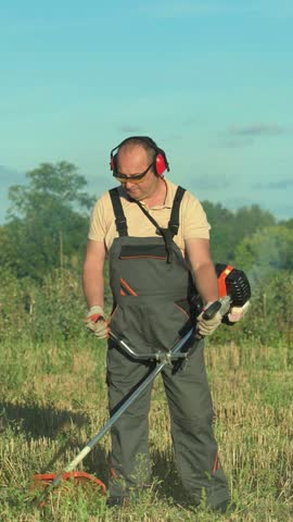 Landscaper in protective earmuffs guides a gas powered brush cutter through tall dry grass, moving the cutting head across an open field with trees and blue sky behind