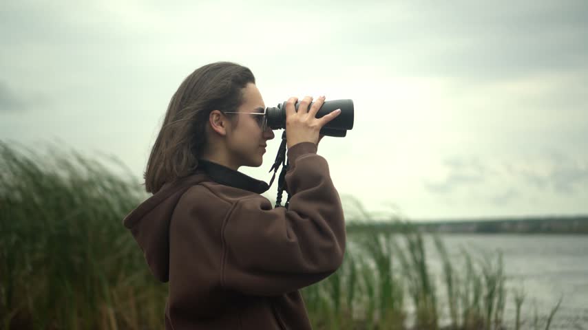 Young Woman in Glasses Looking Through Binoculars at the Opposite Shore of a Lake in Cloudy Weather, Wearing a Brown Tracksuit, Side View, Close-Up