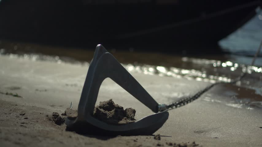 Small Boat Anchor on Chain Near Shore. Anchor Holds a Black Boat in Sunny Weather. Close-Up of the Anchor.