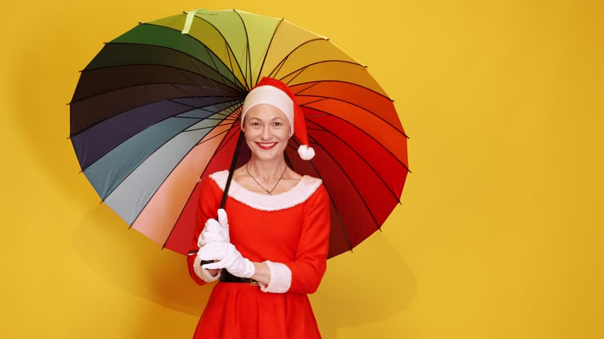 Joyful woman in a santa claus costume happily spinning a colorful rainbow umbrella. Festive and bright concept on a yellow background with copy space