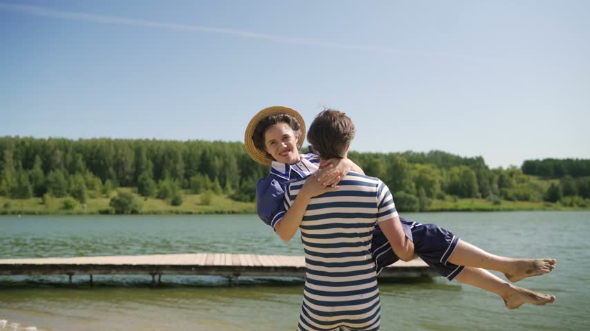 A joyful young couple in retro swimsuits has fun on a lake shore. A man lifts and spins his smiling girlfriend in his arms. A romantic summer vacation scene with a nostalgic feel