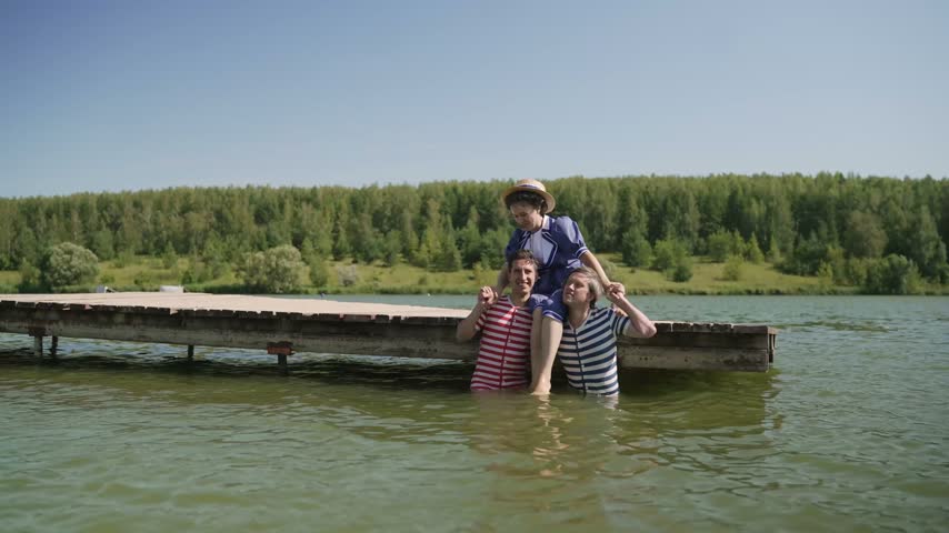 Three young friends in retro bathing suits have fun in a lake on a sunny summer day. Two men lift a smiling woman onto their shoulders, hold the pose, and then playfully lower her into the water