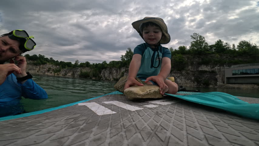 Boy sits on a paddleboard, handles stones and throws them into the water while his father stays alongside at Zakrzowek in Krakow, Poland.