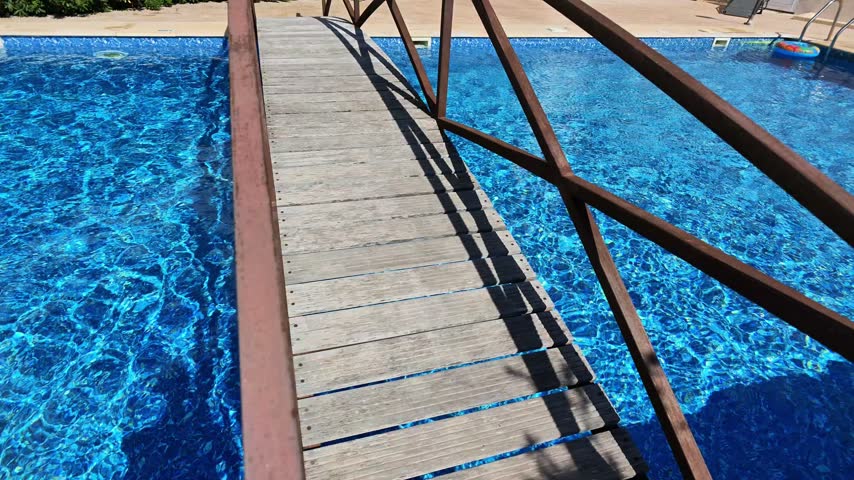 Wooden footbridge crossing a blue swimming pool with metal railings and strong sunlight creating shadows on the walkway