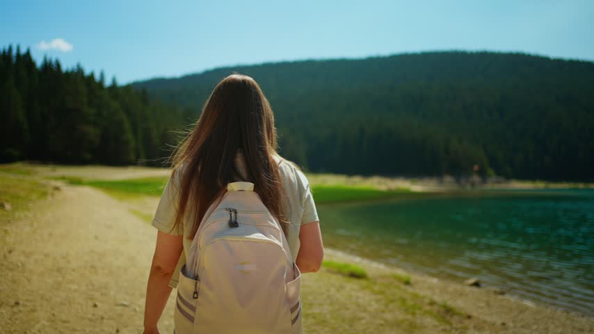 Woman with backpack walking along tranquil lakeside trail toward lush forested hills under bright blue sky on a peaceful summer outdoor adventure