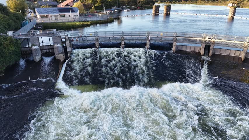 Aerial view of Salmon Weir gates releasing turbulent water into the River Corrib in Galway City, Ireland, with stone structures and the upper river behind.