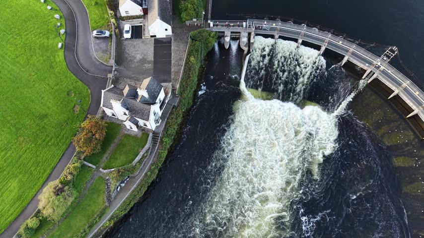 Aerial view of Salmon Weir releasing turbulent water into the River Corrib beside historic buildings and green parkland in Galway City Ireland.