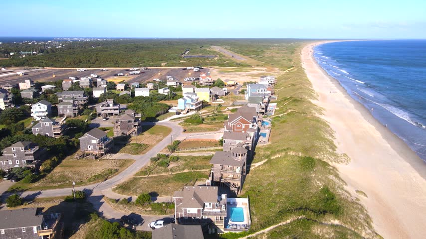 Beachfront homes border grassy dunes beside a long shoreline and breaking ocean waves.
