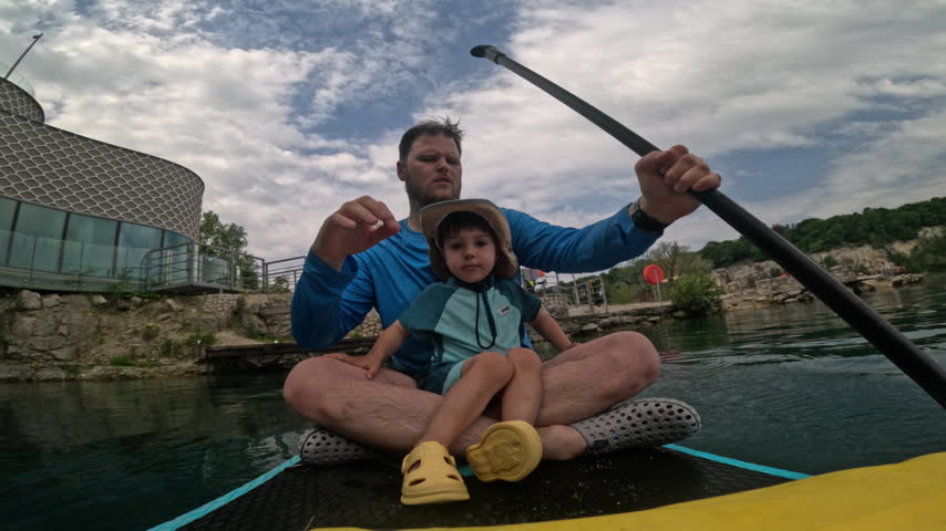 Father paddles a SUP board past a lakeside building while his son sits in front and looks around at Zakrzowek in Krakow, Poland.