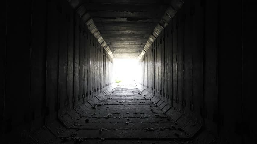 Dark unfinished concrete subway tunnel with repeating structural segments and bright daylight at the end.