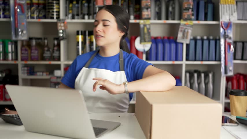 Young latina woman working at the counter of an auto parts store, managing inventory with a laptop