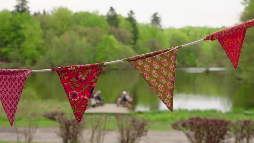 Colorful fabric pennant flags hanging across a lakeside park, with people relaxing by the water in a soft green summer landscape.