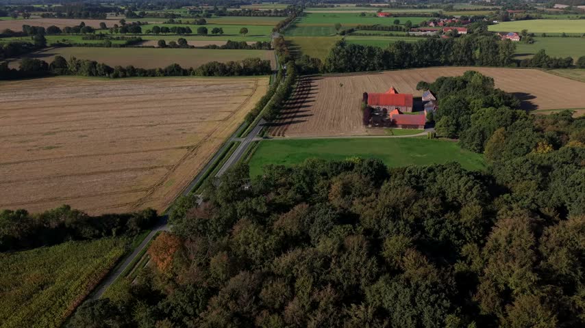 Red-roofed farmhouse sits beside harvested tan field near dense colorful autumn forest, straight tree-lined canal runs parallel to open green lawn beside gravel road. Aerial drone wide shot