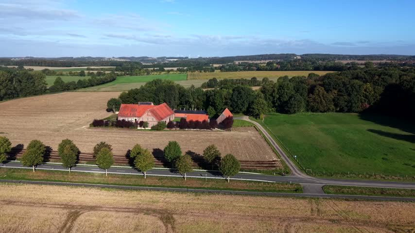 Historic red-roofed farm estate sits within dense trees beside tree-lined road with car in Germany, harvested golden fields stretch toward distant hazy forest under autumn sun. Aerial drone view.
