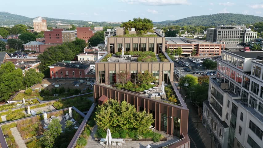 Modern brown brick building with garden terraces in downtown Charlottesville, Virginia, surrounded by older brick blocks, parking lots and the Blue Ridge skyline. Aerial approaching shot