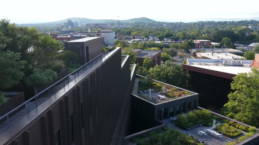 Stepped facade and planted courtyards of a dark brick mixed use building in Charlottesville, Virginia, with downtown rooftops and hazy hills beyond. Aerial approaching shot