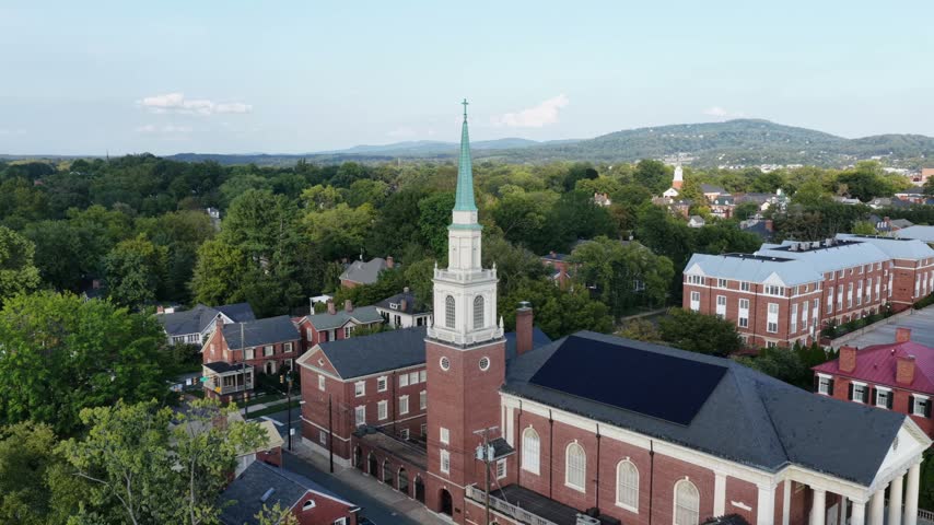 Brick church with a white tower and green copper spire in Charlottesville, Virginia, framed by slate roofs, brick institutional buildings and distant wooded ridges. Aerial approaching shot