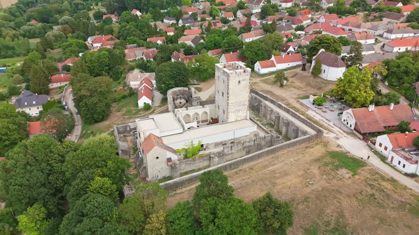Kinizsi Castle in Nagyvázsony, Hungary, medieval fortress with stone walls, tower and moat beside the village center and church, surrounded by greenery in an aerial view.