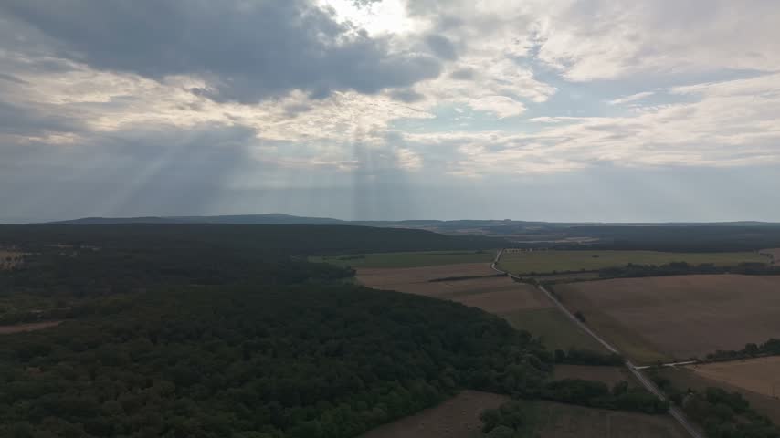 Nagyvázsony, Hungary patchwork farmland, meadows and forested hills under a cloudy summer sky with sun rays across the rural landscape in wide aerial view.
