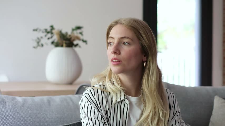 Beautiful young blonde woman with a candid expression listening intently while sitting on a comfortable grey sofa in her living room, eventually breaking into a warm and genuine smile