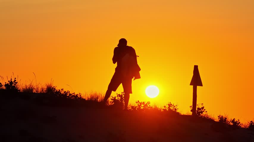 Backlit silhouette of a hiker putting on his backpack and walking along a dune at sunset