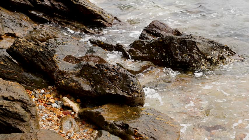 Gentle ocean waves wash over textured dark rocks along a coastal shoreline in Thailand.