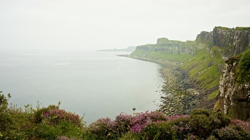 Aerial Drone view looking along dramatic sea cliffs with layered rock and blooming heather. Calm coastal water and rugged terrain create a scenic landscape on Isle of Skye Scotland.