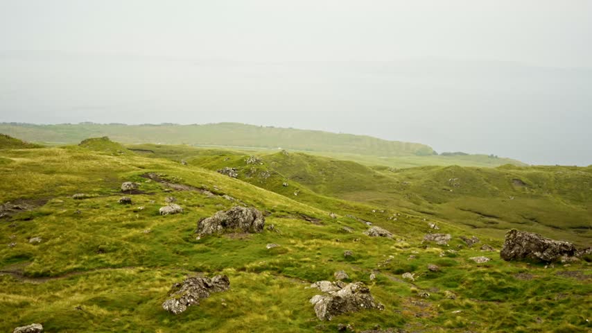 Aerial Drone view panning across lush green moorland with rocky outcrops and rolling hills. Misty conditions create a scenic natural landscape on Isle of Skye Scotland.
