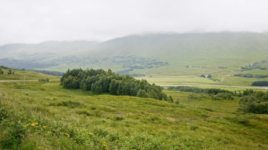 Lush green valley with rolling hills and tree covered slopes beneath misty mountain ranges. Open grassland and distant hills create a scenic countryside landscape in Scotland.