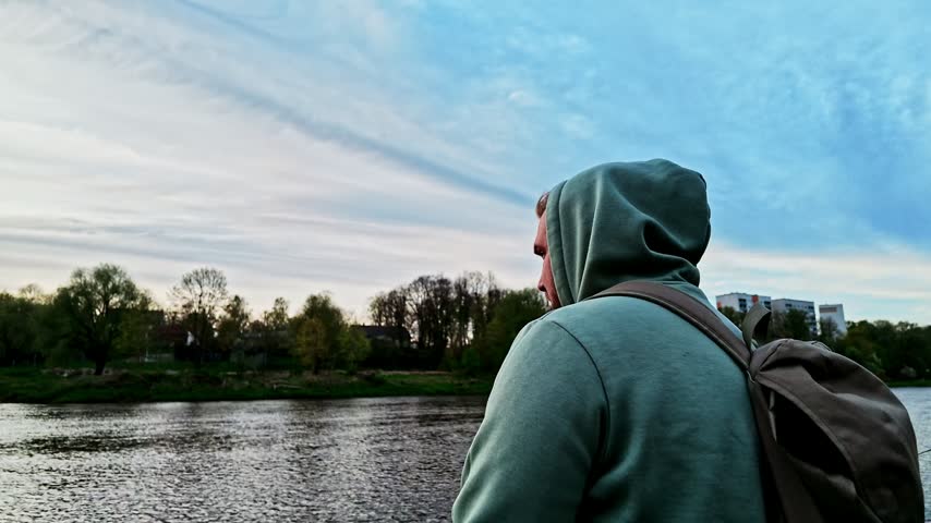 Thoughtful young man wearing a hoodie and backpack looking at the river from the embankment