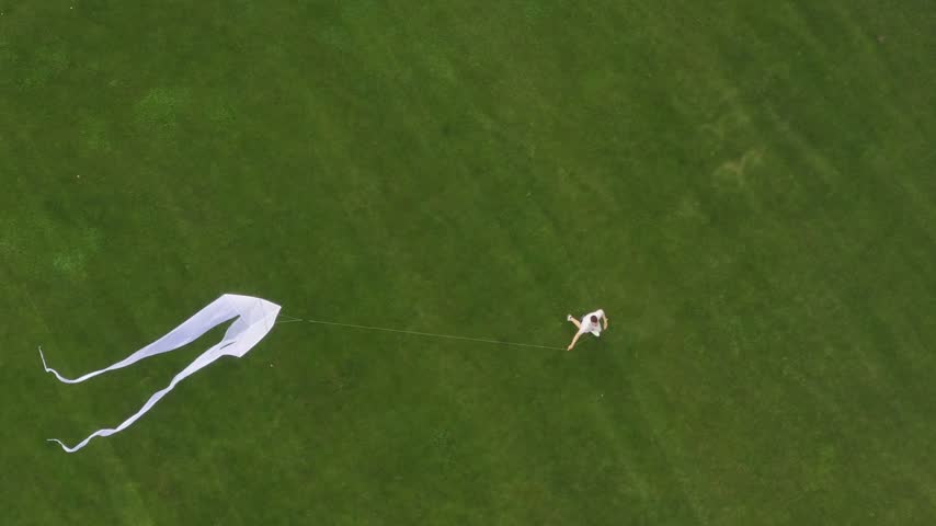 High angle view of a person running across a lush green field while flying a large white kite. Outdoor summer activity and childhood freedom concept