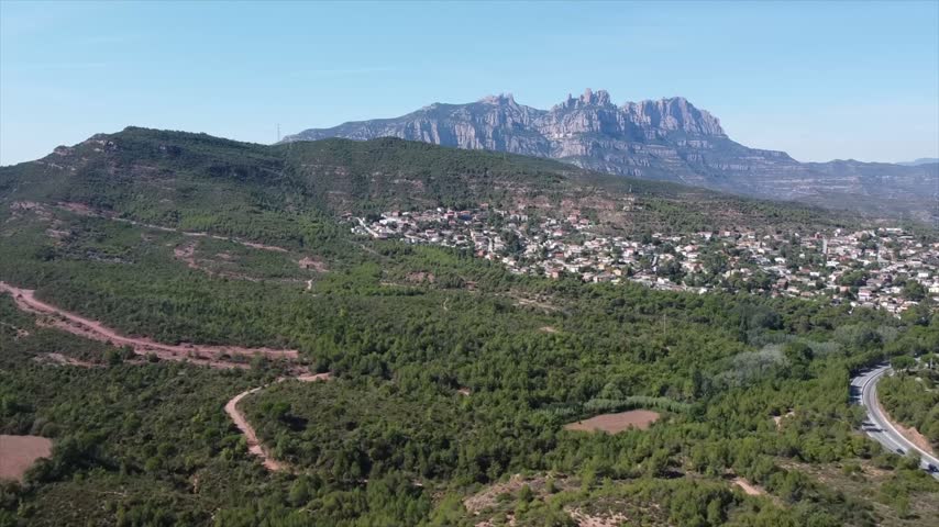 4K drone aerial view of Vacarisses, Catalonia, Spain. The green landscape surrounds the village, with the distinctive rocky peaks of Montserrat rising dramatically in the background.