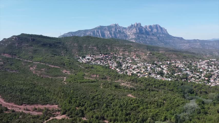 4K drone aerial view of Vacarisses, Catalonia, Spain. The green landscape surrounds the village, with the distinctive rocky peaks of Montserrat rising dramatically in the background.